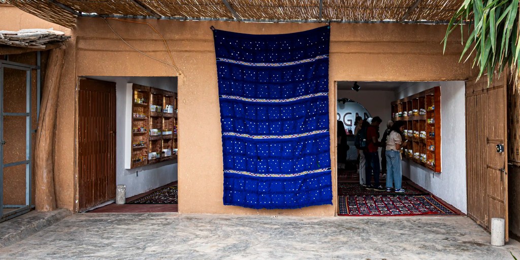 An entrance to a shop with a blue textile hanging above, showcasing shelves filled with products inside. People are visible in the store, with a rustic building and patterned floor covering.