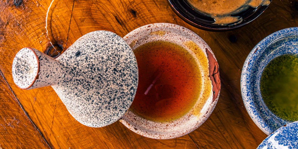 A ceramic dish with a speckled lid containing a rich amber liquid, surrounded by smaller bowls with various sauces, all placed on a wooden surface.