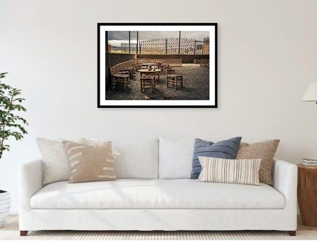 A cozy living room featuring a white sofa adorned with decorative pillows, alongside a wooden side table. A framed picture of an outdoor seating area with wooden chairs and a gravel floor hangs on the wall, providing a warm and inviting ambiance.