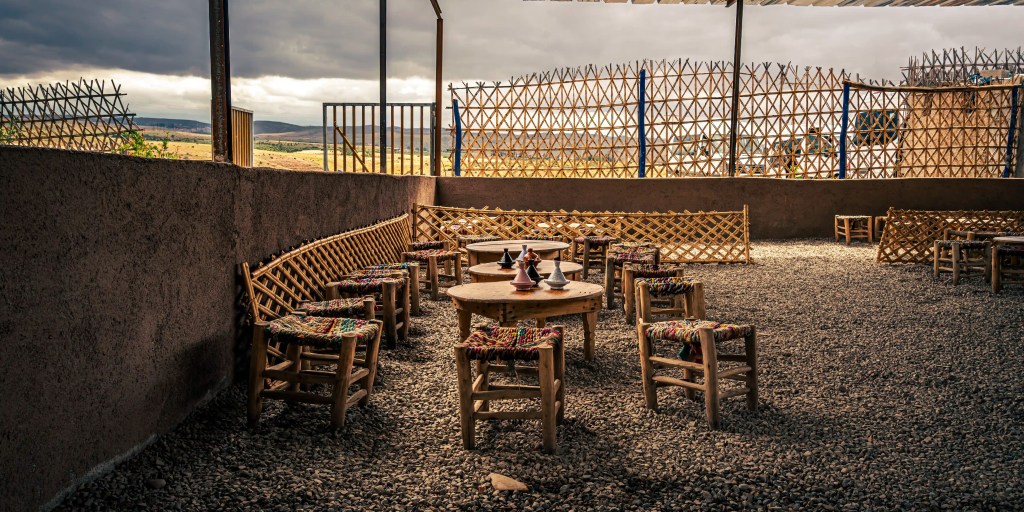 An outdoor seating area with wooden tables and chairs, featuring colorful woven seats. The space is surrounded by a low wall and a wooden lattice fence, with a cloudy sky visible overhead.