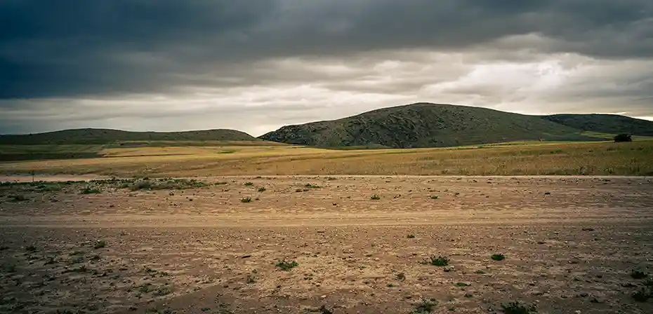 A barren landscape with rolling hills under a cloudy sky, featuring a dirt road and sparse vegetation.