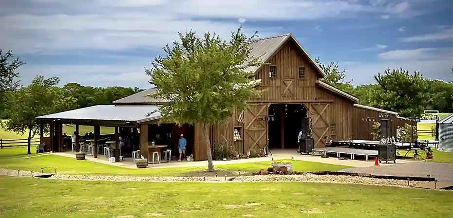 A rustic wooden barn surrounded by grassy fields and trees, featuring an outdoor seating area and a small stage for events.